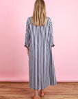A woman with long blonde hair faces away wearing the Xirena Boden Dress—an ankle-length, loose-fit, long-sleeve cotton poplin shirtdress with vertical black and white stripes—and orange sandals, against a pink background and wooden floor.