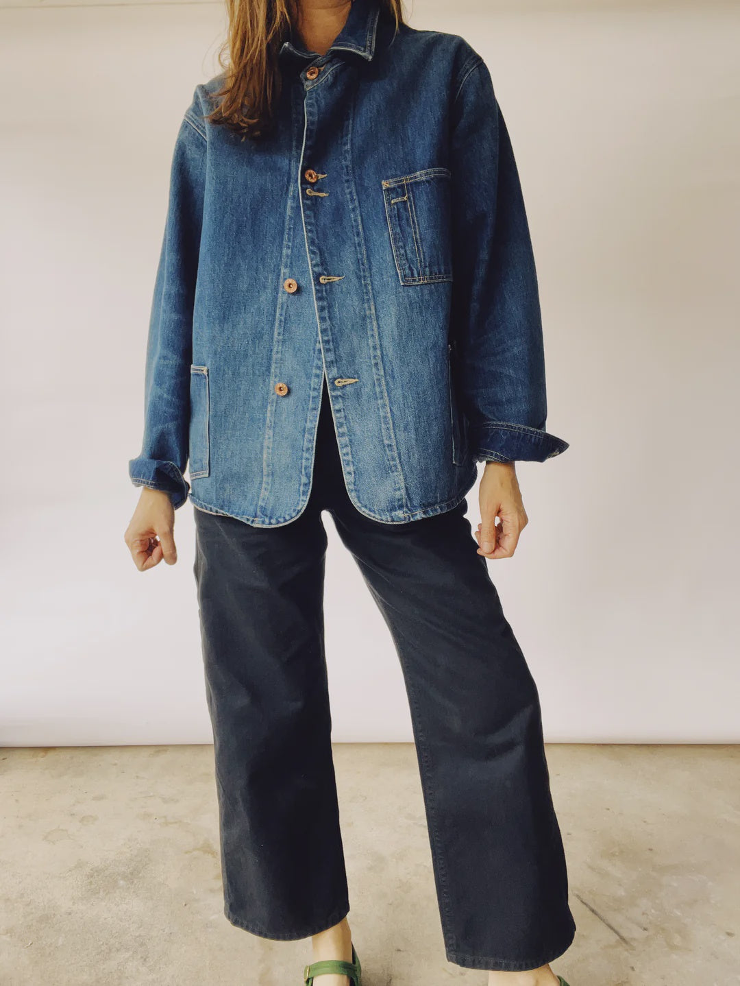 A person wearing The Barn Coat by Hey Gang, an oversized unisex Japanese denim jacket featuring large front pockets and brown buttons, paired with black wide-leg pants, stands against a plain background. Their hand is slightly raised.