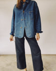 A person wearing The Barn Coat by Hey Gang, an oversized unisex Japanese denim jacket featuring large front pockets and brown buttons, paired with black wide-leg pants, stands against a plain background. Their hand is slightly raised.