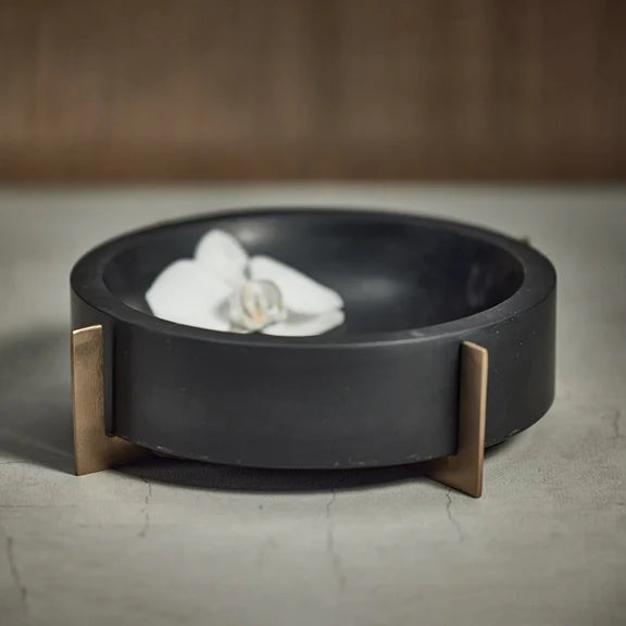 A Zodax Black Marble Bowl with a gold metal base holds a white orchid, serving as a decorative catch-all on a textured light gray surface against a blurred brown background.