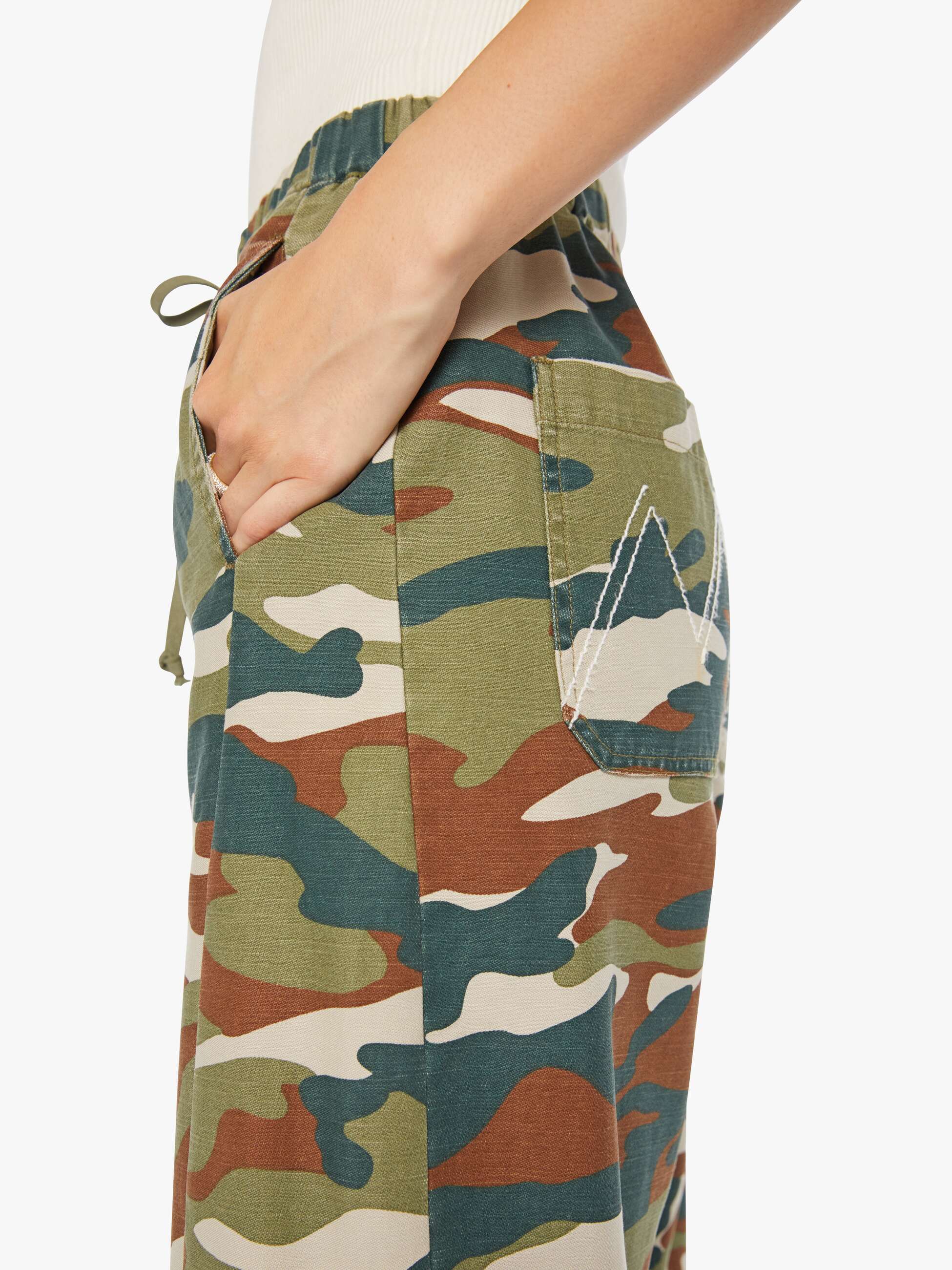 A person wears The Drawn Half-Pipe Ankle by Mother—high-waisted cotton pants in green, brown, and beige camo with a drawstring waist, white stitching on the back pocket, and a hand resting in the pocket.