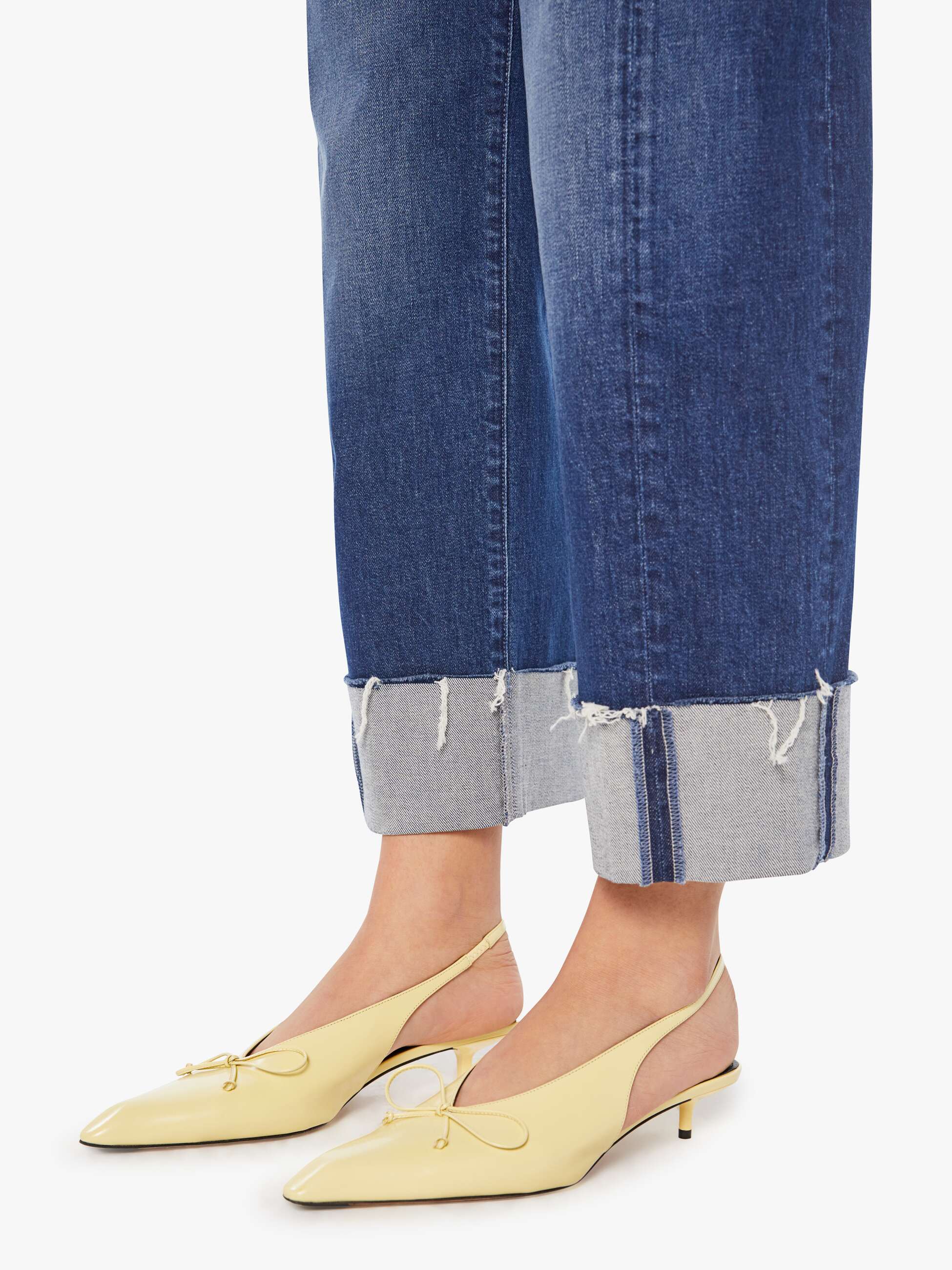 A person wears Mother’s The Half-Pipe Ankle Cuff Fray high-waisted barrel jeans and pale yellow slingback kitten heels with a bow, standing against a white background.