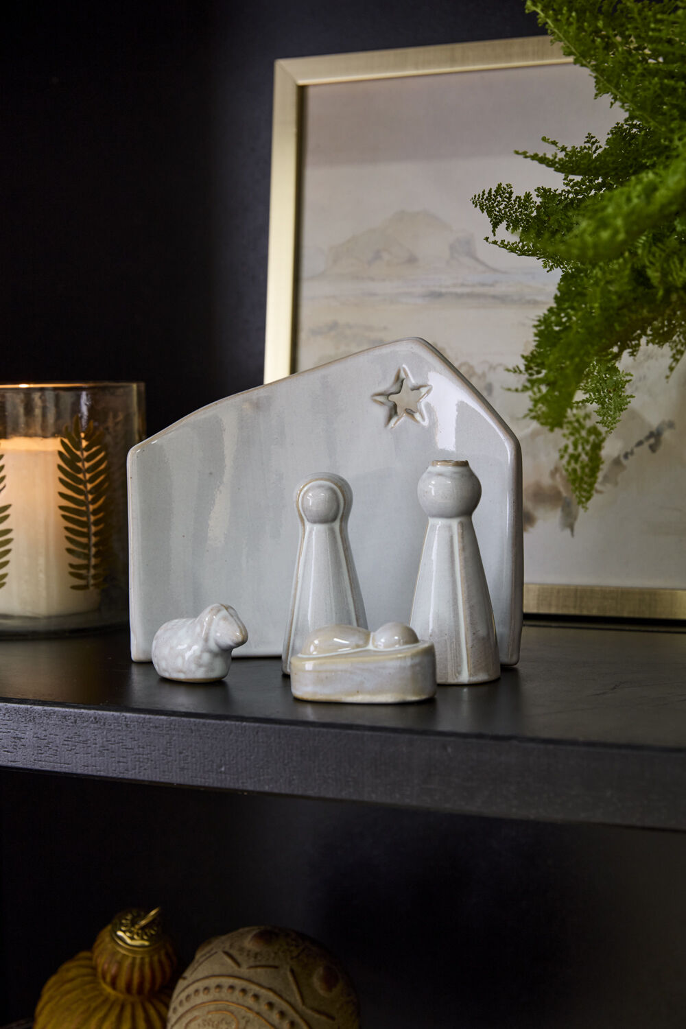 The Claro Nativity Set by Accent Decor, a small white ceramic display featuring Mary, Joseph, baby Jesus, a sheep, and a star, sits on a black shelf as a festive centerpiece beside a candle, green plant, and framed photo.