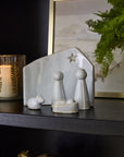 The Claro Nativity Set by Accent Decor, a small white ceramic display featuring Mary, Joseph, baby Jesus, a sheep, and a star, sits on a black shelf as a festive centerpiece beside a candle, green plant, and framed photo.