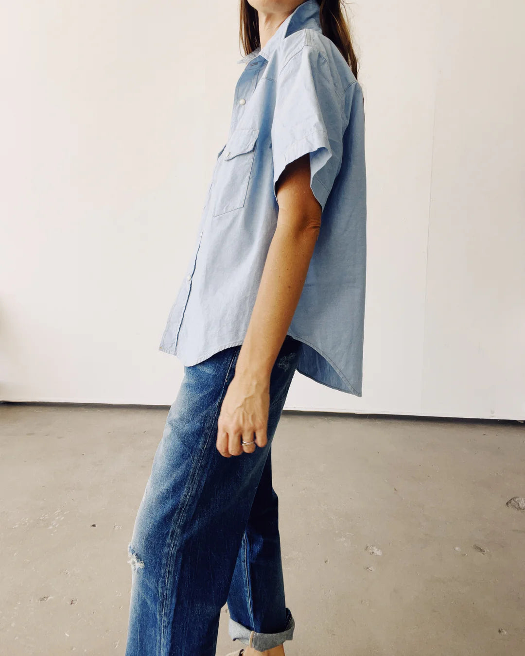 A person wearing The Western Shirt by Hey Gang, styled as a light blue oversized shirt, paired with blue jeans stands in a minimalist room with a concrete floor and plain white walls. The focus is on their casual outfit.