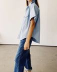 A person wearing The Western Shirt by Hey Gang, styled as a light blue oversized shirt, paired with blue jeans stands in a minimalist room with a concrete floor and plain white walls. The focus is on their casual outfit.