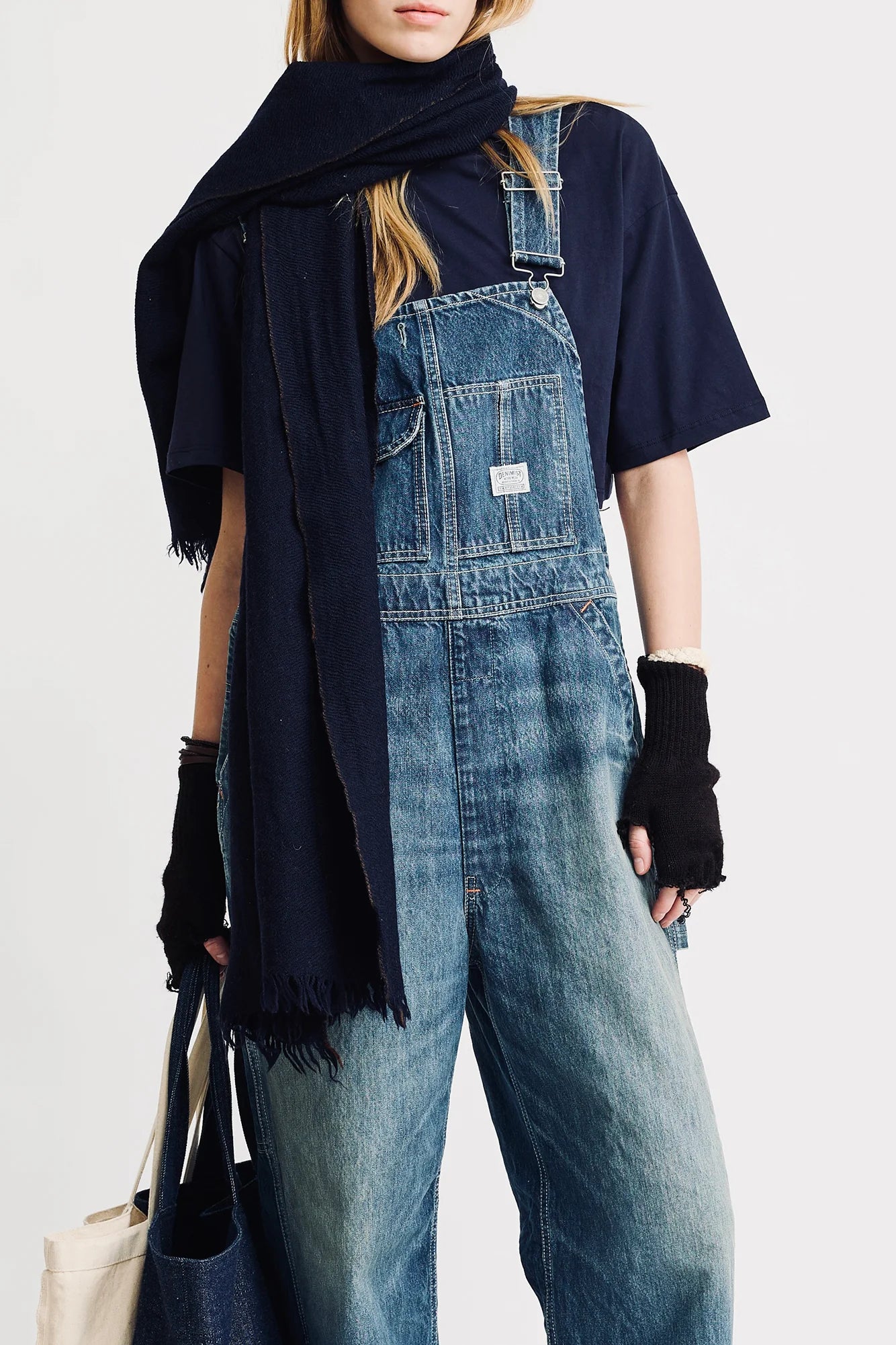 A person in Denimist SLOUCH BIB OVERALL, with a utility pocket, navy tee, dark scarf, black fingerless gloves, and holding a tote bag stands against a plain light background.