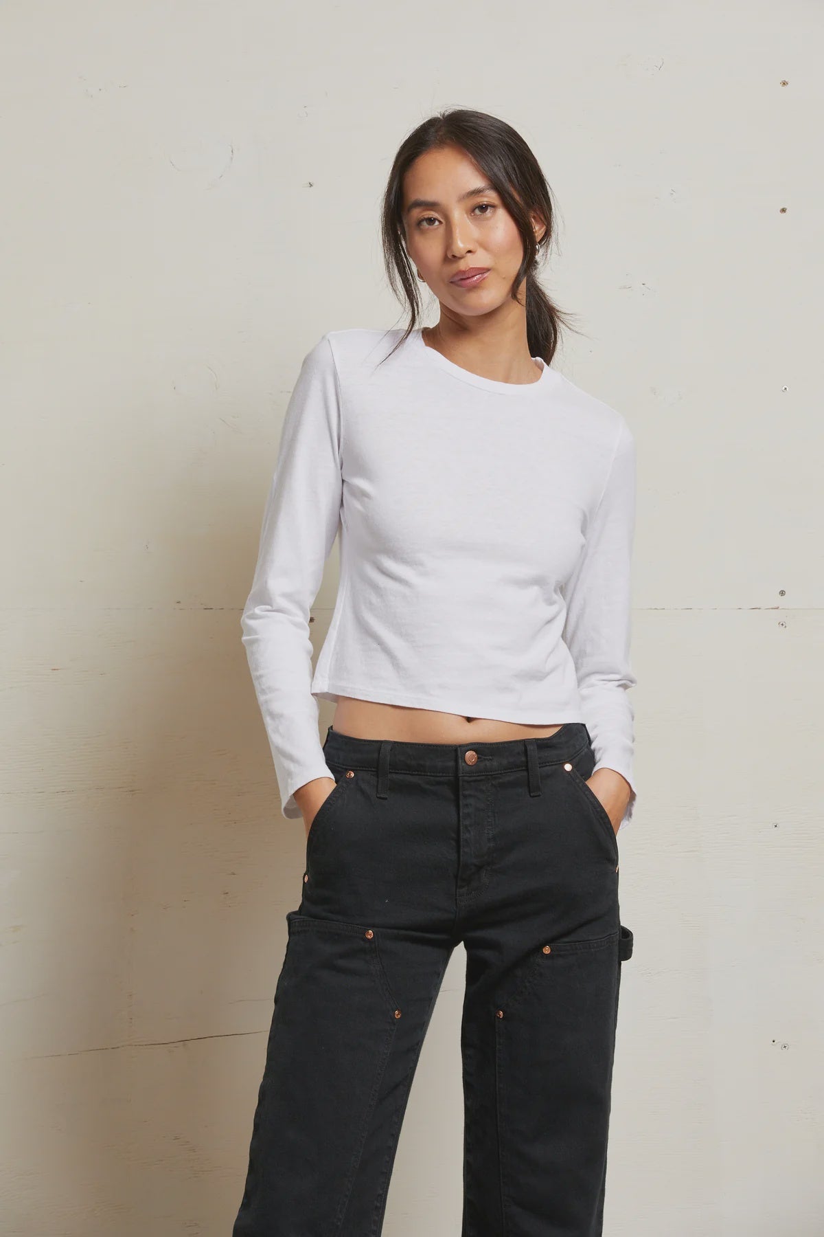 A woman with long dark hair in a low ponytail wears the Perfectwhitetee Jamie LS Crew and black pants, standing against a plain light-colored wall with her hands in her pockets.