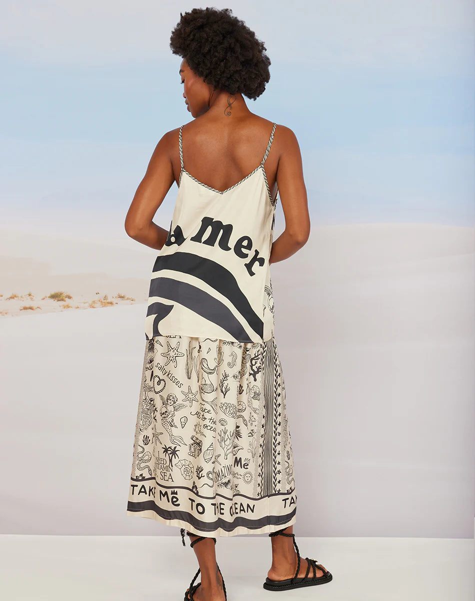 A woman with short curly hair, barefoot and facing away, wears the Me369 Elizabeth Midi Skirt—a vintage-style piece featuring black graphic designs and the text "TAKE ME TO THE OCEAN.