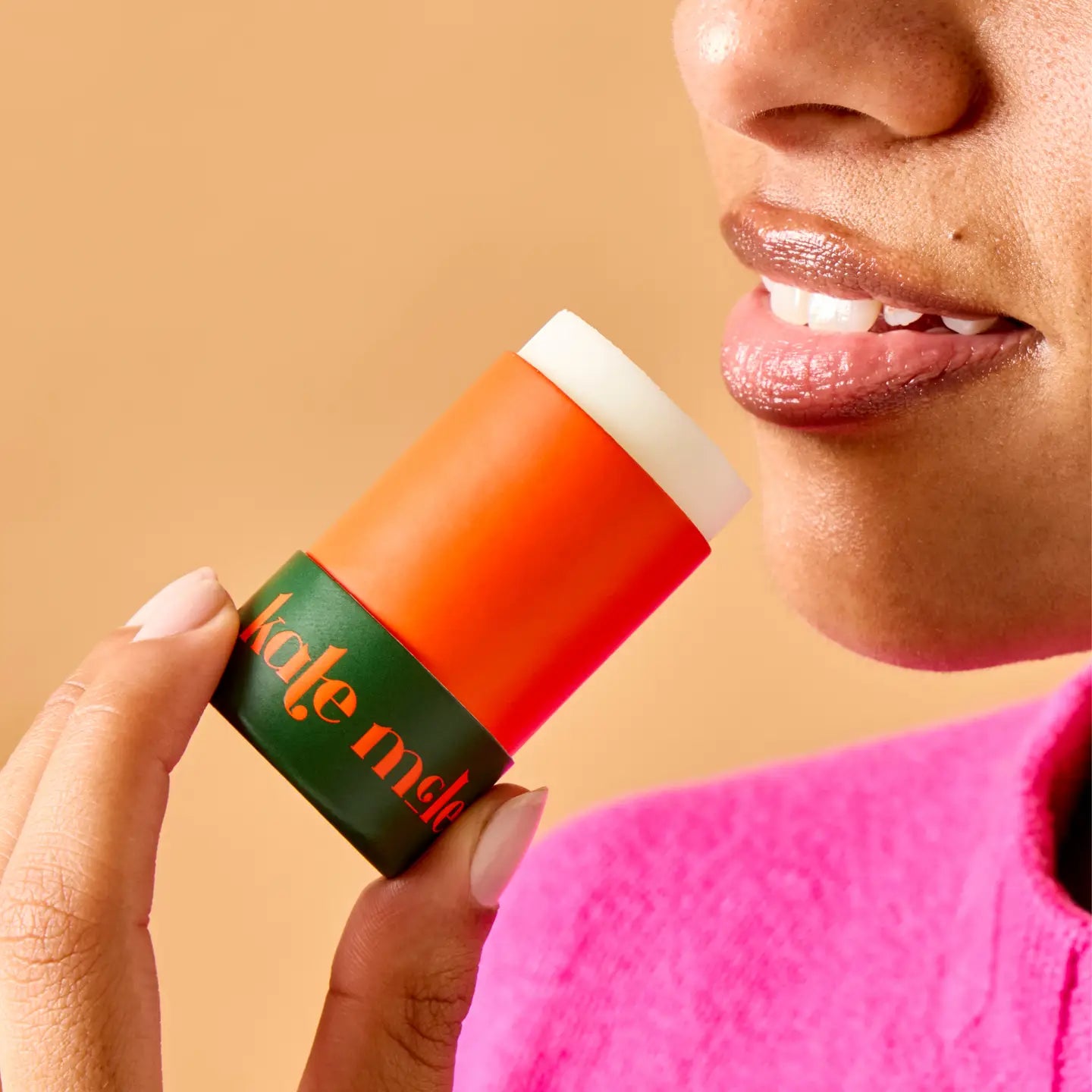 A woman in a bright pink top gently smiles while holding Faire's "The Everywhere Stick" in orange and green close to her lips against a soft brown backdrop, highlighting its wax-free, true hydration formula as it enhances her glossy lips.