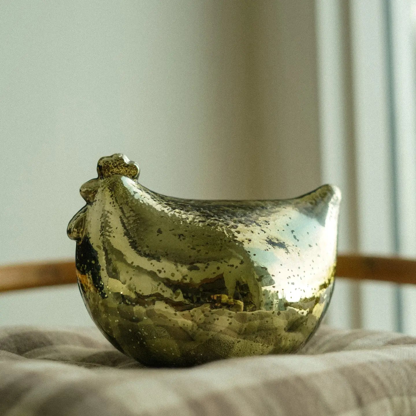 The Indaba Mother Hen Glass Statuette, a gleaming gold-toned decor piece, rests on beige fabric with a softly blurred backdrop that includes a window and wooden furnishings.