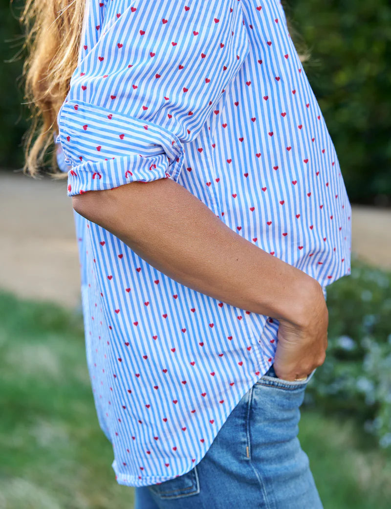 A person with long hair stands outdoors in a relaxed-fit Frank & Eileen Eileen shirt with blue and white stripes and small red hearts, sleeves rolled up, paired with Italian cotton blue jeans, hands in pockets, against a blurred green backdrop.