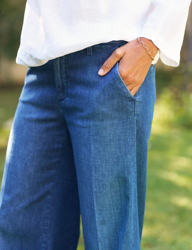 A person stands outdoors with one hand in their pocket, wearing a white long-sleeve shirt, Frank &amp; Eileen NYC Trouser jeans in blue denim, and a gold bracelet on their wrist.