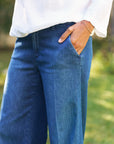A person stands outdoors with one hand in their pocket, wearing a white long-sleeve shirt, Frank & Eileen NYC Trouser jeans in blue denim, and a gold bracelet on their wrist.