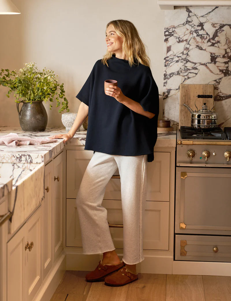 A woman in a navy top and Frank & Eileen's Bella Sweatpants stands smiling in a stylish kitchen with marble countertops, a decorative vase with flowers, and stainless steel appliances. She wears brown sandals while holding a cup.