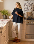 A woman in a navy top and Frank & Eileen's Bella Sweatpants stands smiling in a stylish kitchen with marble countertops, a decorative vase with flowers, and stainless steel appliances. She wears brown sandals while holding a cup.