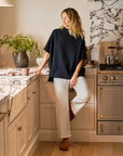 A person in Frank & Eileen's Bella Sweatpant stands in a cozy kitchen, leaning against a marble countertop. Plants adorn a vase, a kettle sits on the stove, and soft lighting creates warmth.