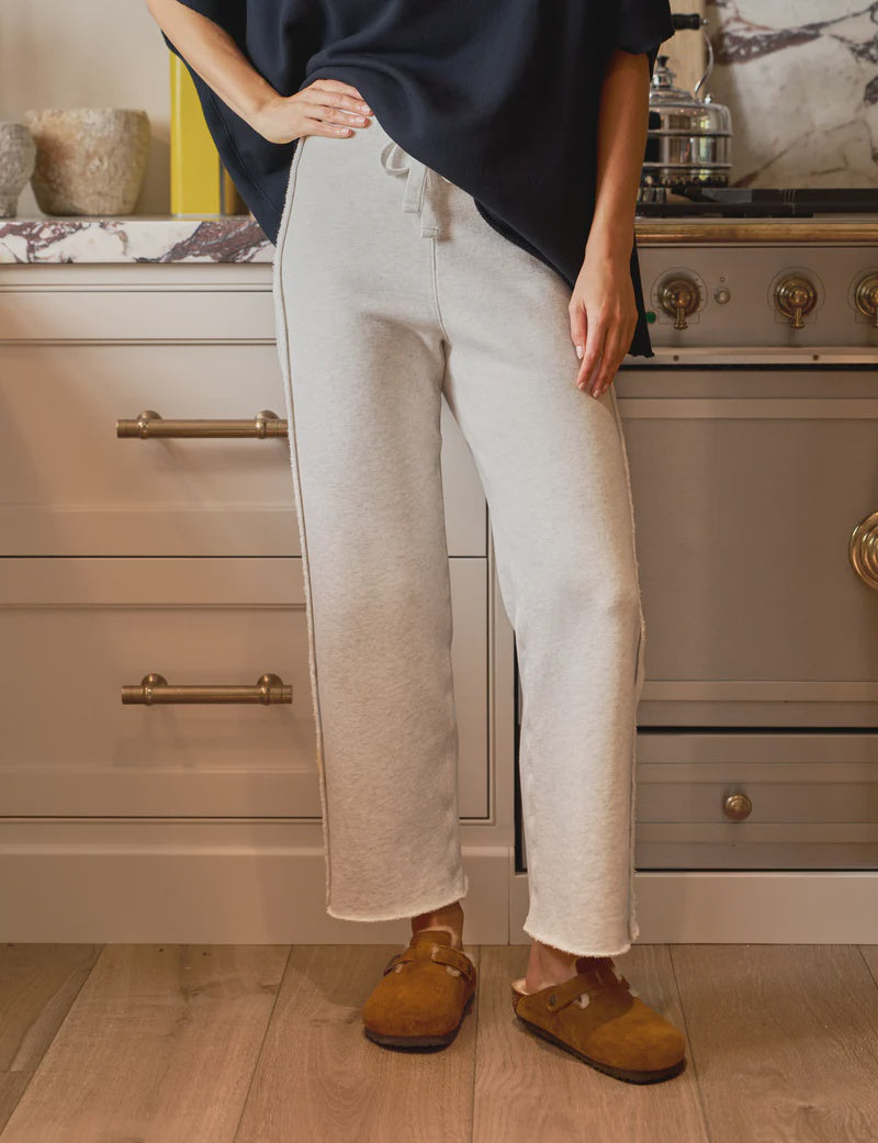 A person in light gray, high-rise Bella Sweatpants by Frank & Eileen and brown slippers stands in a kitchen with cabinets, a decorated countertop, and part of a stove. These 100% cotton sweatpants combine comfort with style.