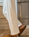 A close-up shows a person wearing Frank & Eileen's Bella Sweatpants paired with brown suede mule slippers, walking on a wooden kitchen floor with stainless steel appliances.