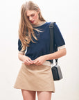 A woman with long blonde hair wears the Many Contrast T-Shirt by Jumper 1 2 3 4/CR2, paired with a beige mini skirt and black shoulder bag. She looks down, hands near her waist, against a plain light background.