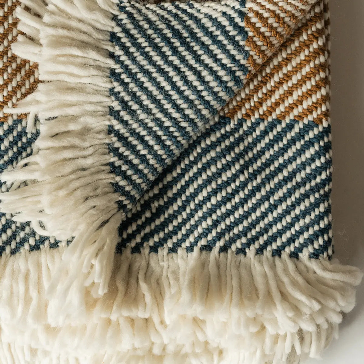 A close-up of the Faire Chunky Twill Throw, folded to reveal its blue, white, and brown herringbone knit pattern with thick white fringe edging.
