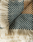 A close-up of the Faire Chunky Twill Throw, folded to reveal its blue, white, and brown herringbone knit pattern with thick white fringe edging.