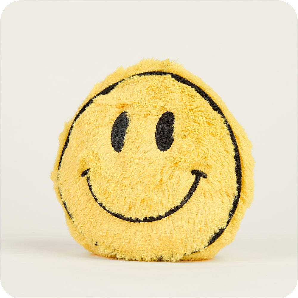 The Smiley Face Warmies Junior by Faire is a round, yellow plush toy with a fuzzy texture, black smiley face, and black outlines, designed for sensory comfort against a plain light background.