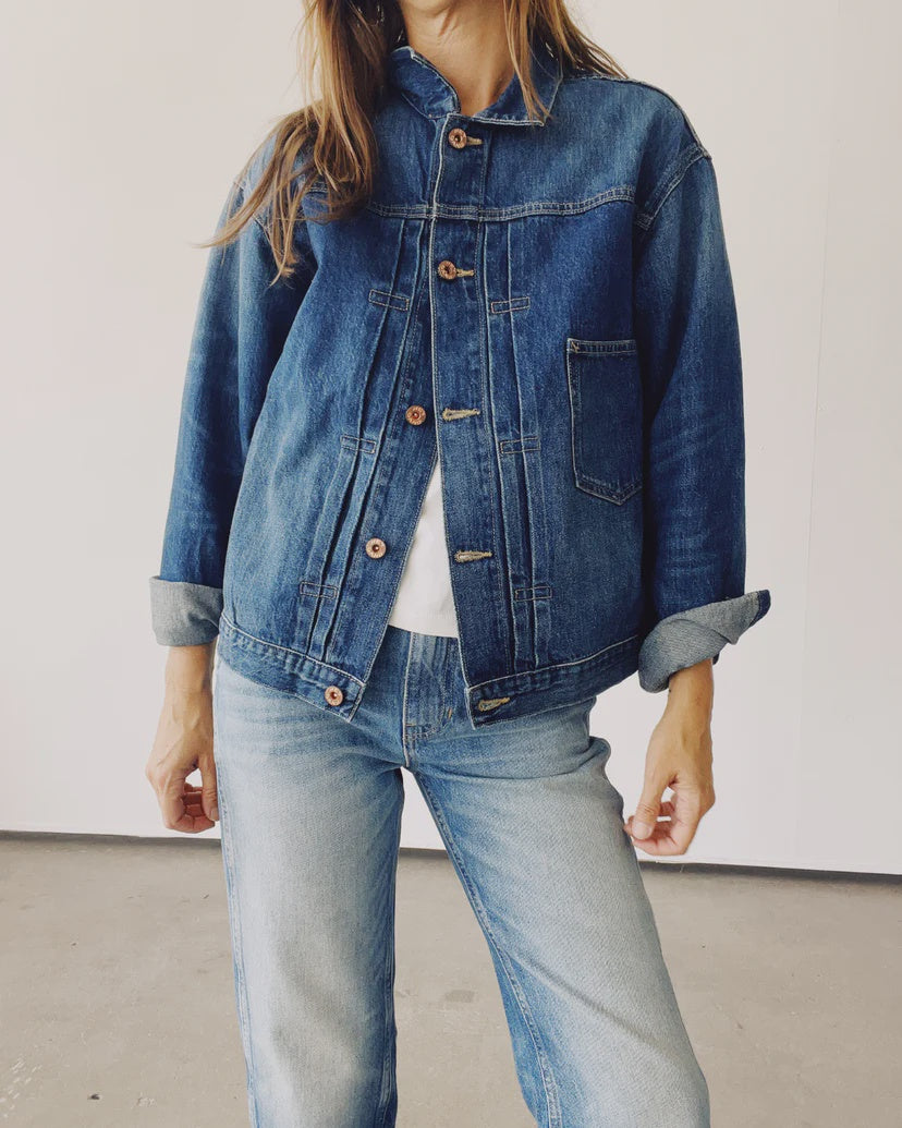 A person dressed in "The Denim Jacket" by Hey Gang and a pair of jeans stands against a plain background. The jacket is buttoned, showcasing a white shirt underneath, with the sleeves stylishly rolled up.