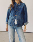 A person dressed in "The Denim Jacket" by Hey Gang and a pair of jeans stands against a plain background. The jacket is buttoned, showcasing a white shirt underneath, with the sleeves stylishly rolled up.