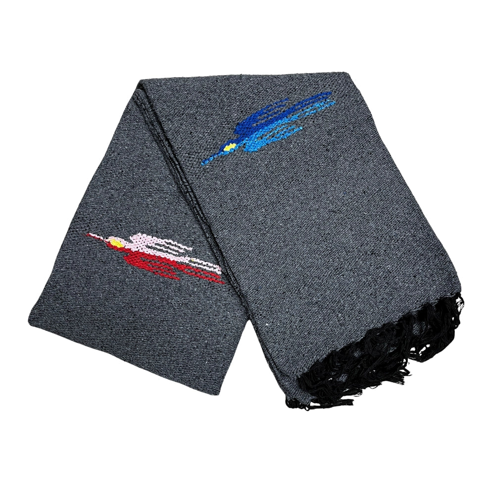The Charcoal Thunderbird Baja Blanket by Faire is a gray woven piece with black fringe and two embroidered fish—one blue, one red and pink—blending artisanal design with the cozy feel of a handmade blanket.