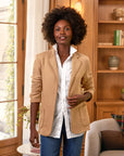 A woman with curly hair wears the Frank & Eileen Dublin Knit Blazer over a white shirt and blue jeans, smiling indoors in a bright room with wooden shelves, a plant, and a window in the background.