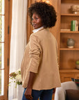 A woman with curly hair wears the Frank & Eileen Dublin Knit Blazer in a tailored fit over a white shirt, glancing over her shoulder in a cozy room with wooden shelves, books, and a vase of flowers on the nearby table.