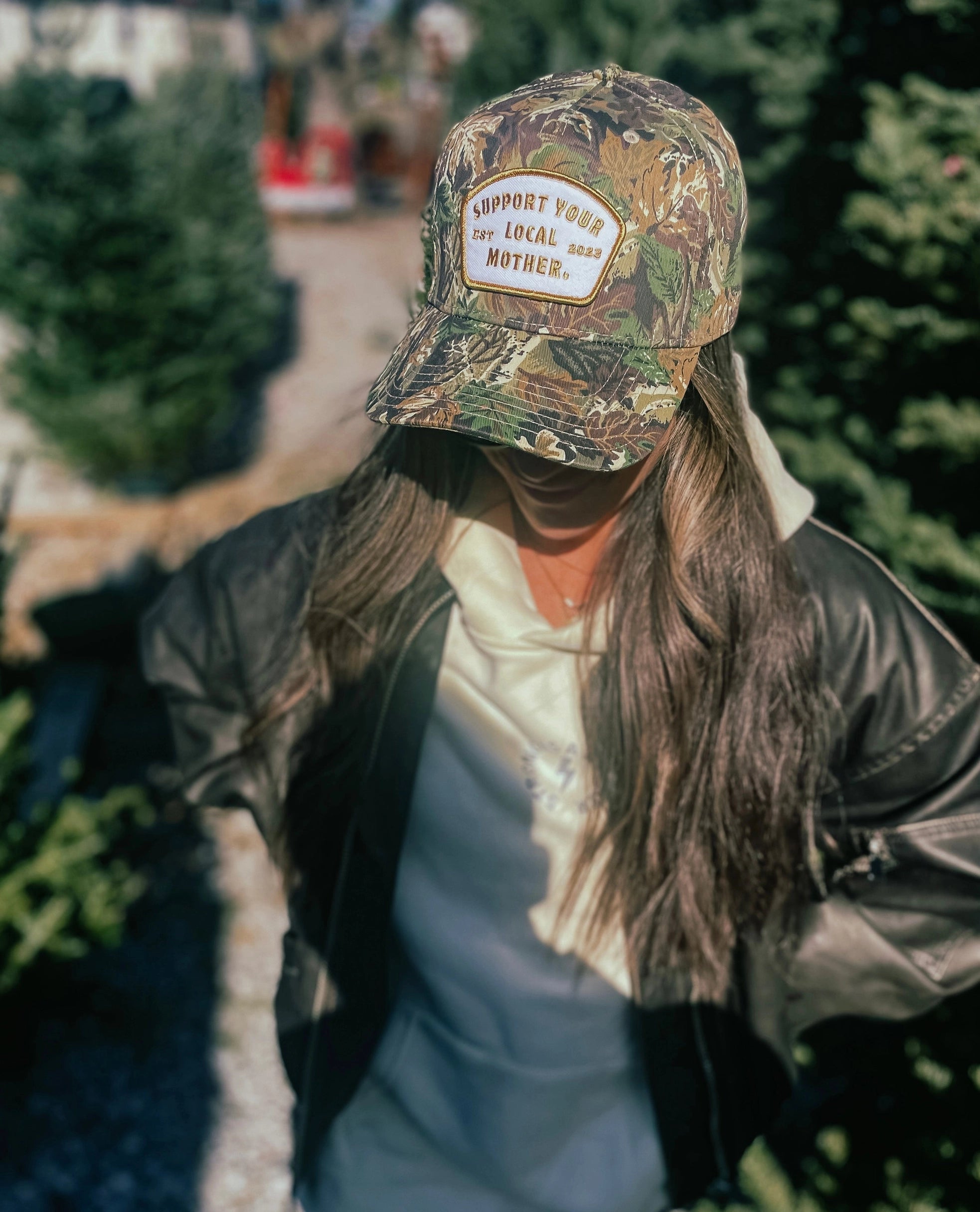 Outdoors near evergreens, someone in a cream hoodie, dark jacket, and Faire’s Support Your Local Mother camo canvas hat stands with the brim shadowing their face, bringing a sense of intrigue to the serene scene.