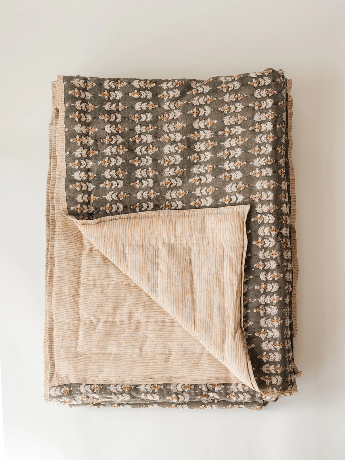 The Faire Blossom Block Print Quilt is folded on a white background, showing its floral gray and white pattern on one side and light beige stripes on the other. A flipped corner reveals the bohemian bedding style featured on both sides.