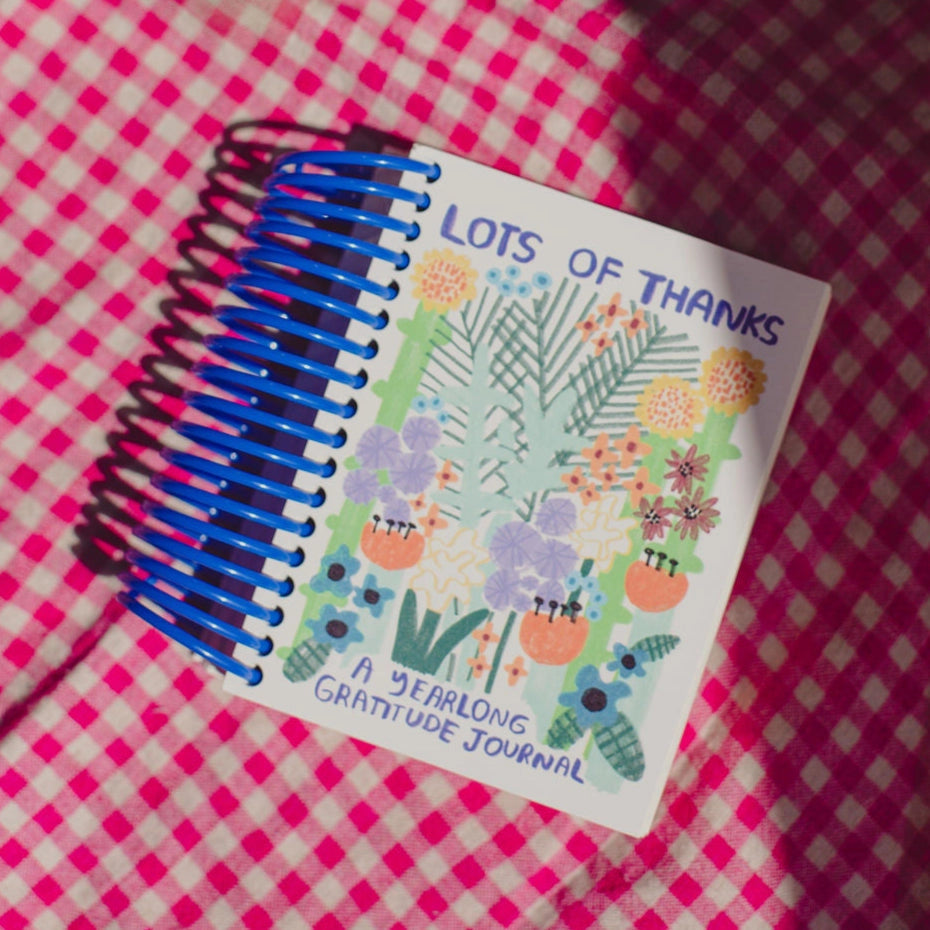 A spiral-bound Faire Gratitude Journal with a colorful floral cover, made in the USA, rests on pink and white checkered fabric.