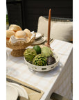 A table set for a meal with a white tablecloth features the Indaba Cypress Fruit Bowl L filled with artichokes and avocados, alongside a basket of bread rolls, beige candle, and cushioned seating—an ideal kitchen accessory for any countertop.