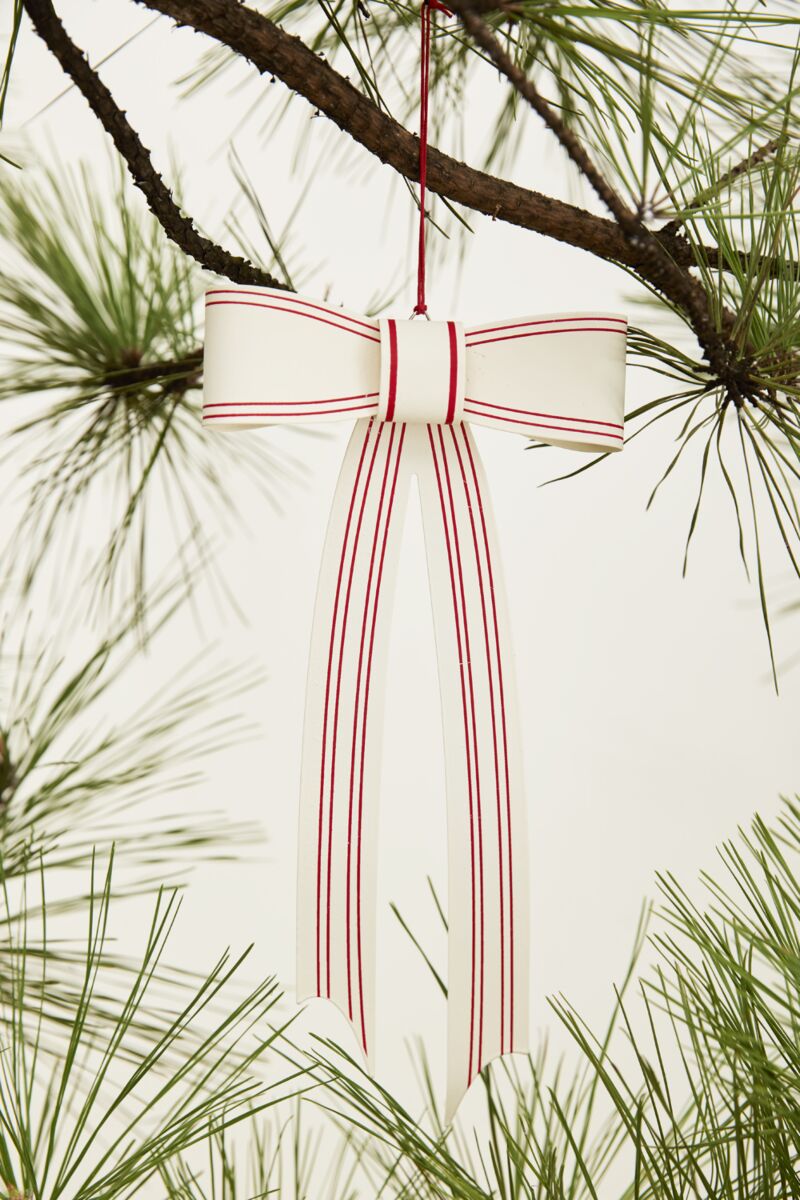 The Candace Ornament 6x12 by Accent Decor, a white ribbon with red stripes tied in a bow, hangs from a pine branch as a charming Christmas tree decoration amid green needles and a plain light background.