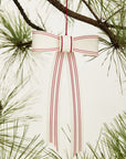 The Candace Ornament 6x12 by Accent Decor, a white ribbon with red stripes tied in a bow, hangs from a pine branch as a charming Christmas tree decoration amid green needles and a plain light background.