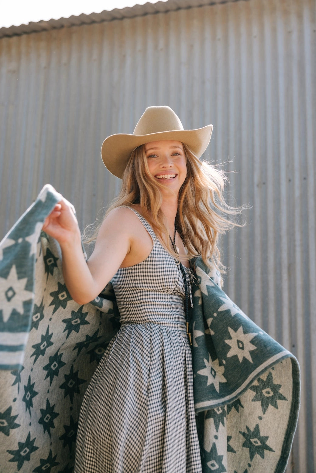 A smiling woman with long, wavy hair wears a gingham dress and tan cowboy hat, draping the Faire Big Sky Lightweight Blanket around her shoulders as she stands outdoors before a corrugated metal wall.
