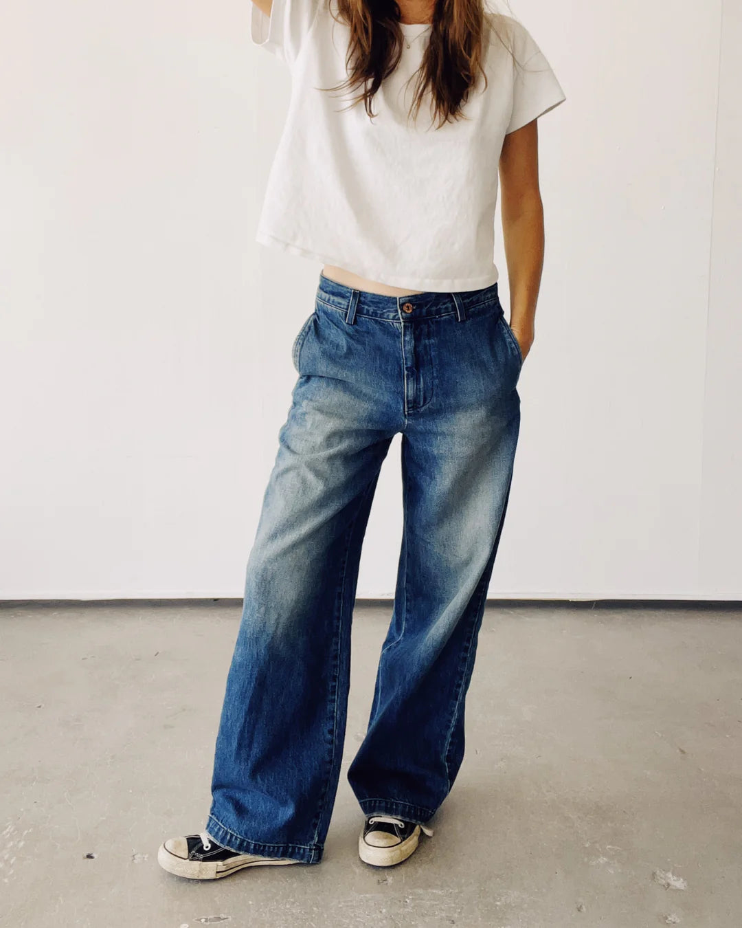 A person stands against a plain white backdrop, wearing a white t-shirt and The Buckle Back Trouser by Hey Gang—slouchy-fit blue jeans made in Los Angeles from cotton denim, paired with black sneakers. One hand is partly raised while the other rests casually in their pocket. The jeans exhibit a faded texture, enhancing their relaxed vibe.