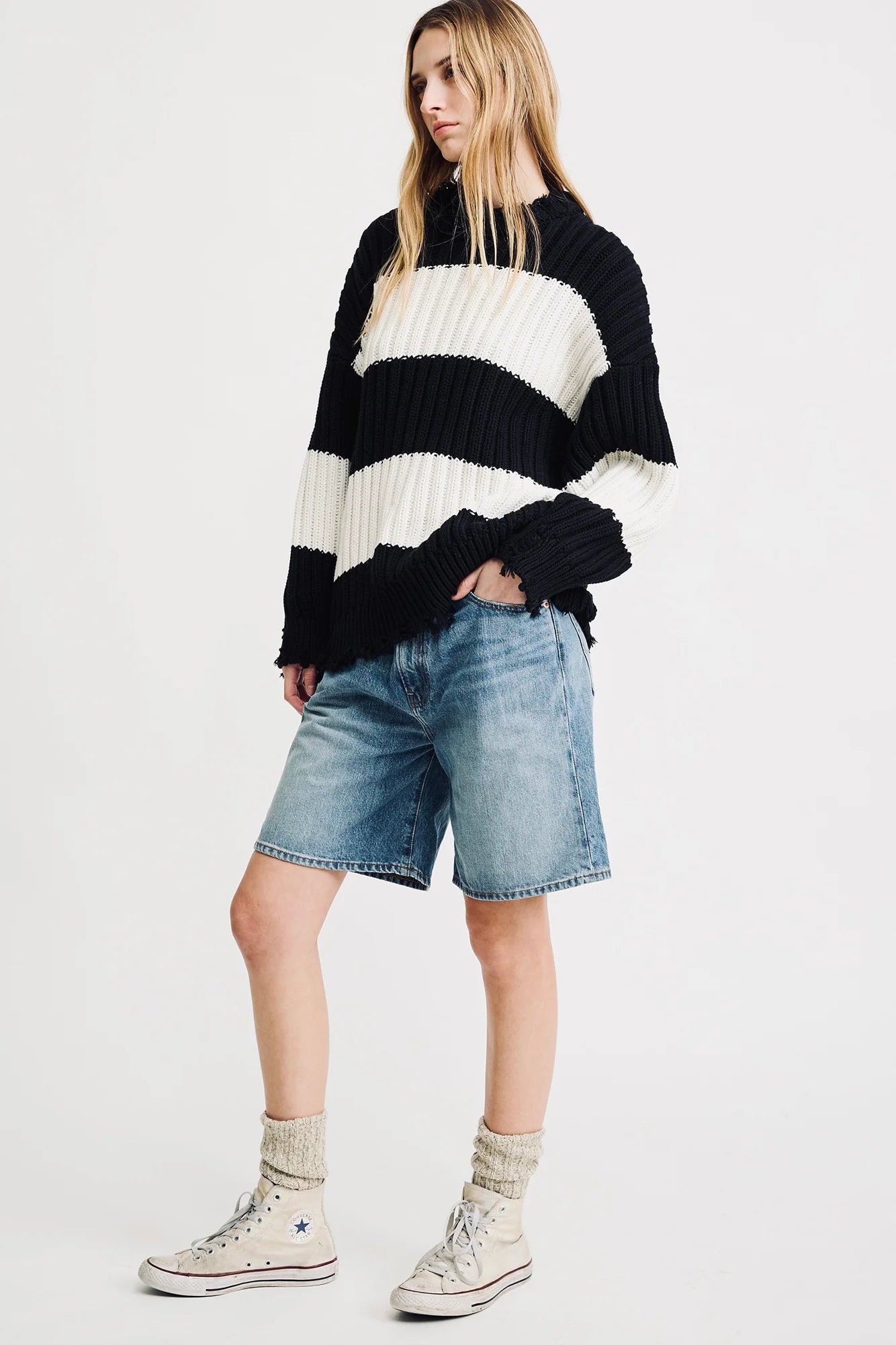 A person with long, loose hair stands against a plain background wearing the Denimist OVERSIZED RIBBED SWEATER with black and white stripes, paired with knee-length denim shorts, light socks, and white high-top sneakers.