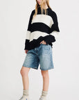 A person with long, loose hair stands against a plain background wearing the Denimist OVERSIZED RIBBED SWEATER with black and white stripes, paired with knee-length denim shorts, light socks, and white high-top sneakers.