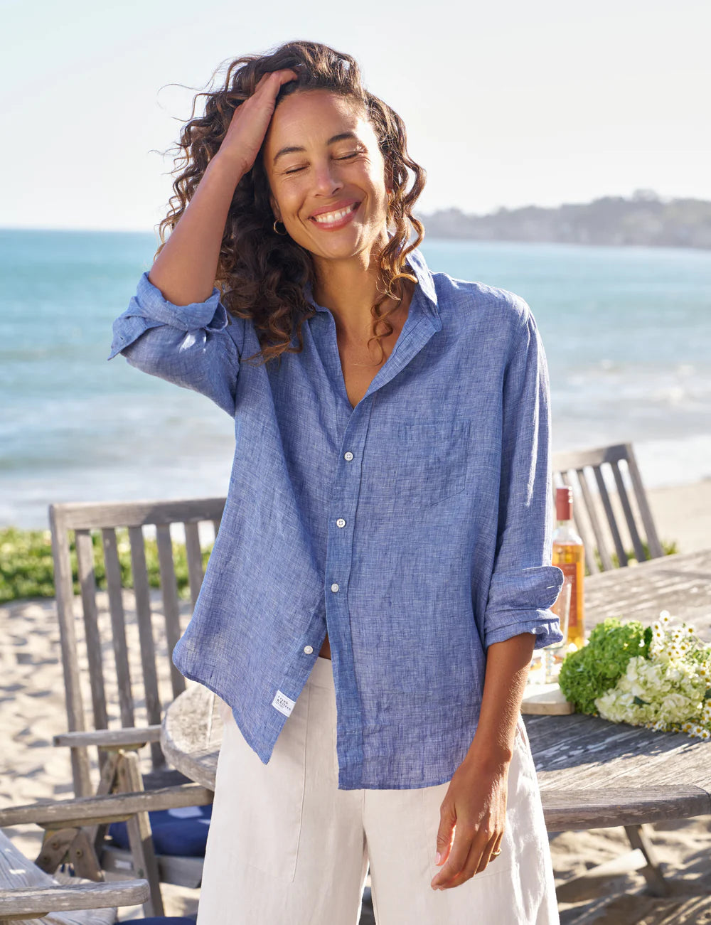 A woman with curly hair smiles with her eyes closed, wearing the Frank & Eileen Eileen 26 relaxed fit 100% cotton blue shirt and white pants, standing on a beachside patio with wooden chairs, a table, and the ocean in the background.