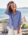 A woman with curly hair smiles with her eyes closed, wearing the Frank & Eileen Eileen 26 relaxed fit 100% cotton blue shirt and white pants, standing on a beachside patio with wooden chairs, a table, and the ocean in the background.