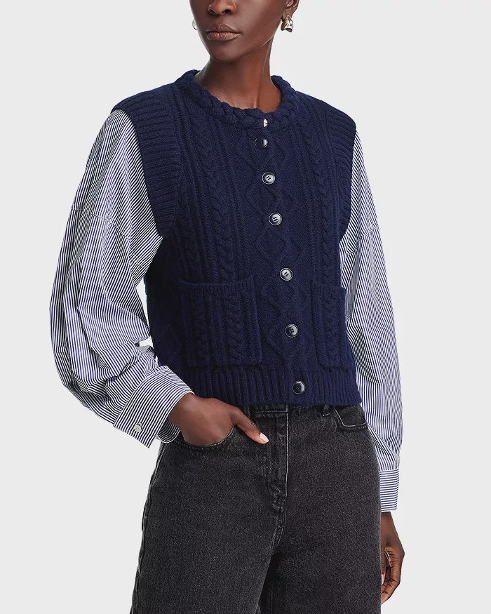 A person is sporting a stylish Caleb Cable Sweater from Sea New York, featuring a blue cable-knit buttoned vest over a blue and white blouse with voluminous cotton poplin sleeves. Complementing the look are sleek black jeans.