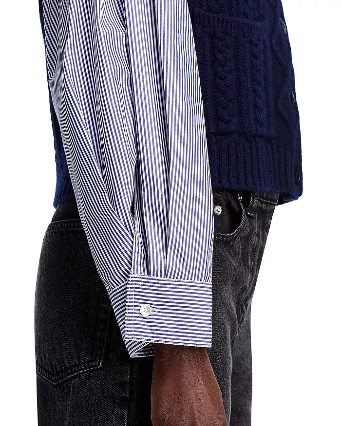 A person is depicted from the side wearing a blue and white striped shirt featuring button front details and cotton poplin sleeves. Over this, they are sporting the Caleb Cable Sweater from Sea New York, a dark blue cable knit sweater vest. Their arm hangs down casually, paired with sleek black jeans.