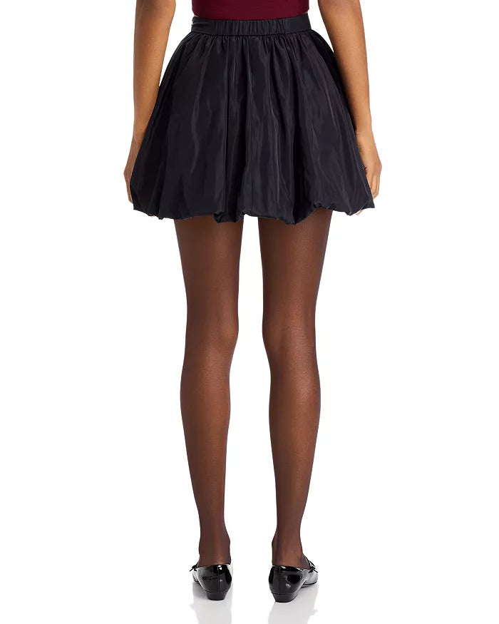 A person stands with their back to the viewer, wearing the Rhode Blanca Skirt, a black pleated mini skirt that suggests an A-line silhouette. Sheer black tights and ballet flats finish off the ensemble, while a deep red top subtly stands out against the plain white background.