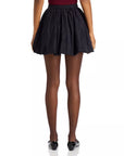 A person stands with their back to the viewer, wearing the Rhode Blanca Skirt, a black pleated mini skirt that suggests an A-line silhouette. Sheer black tights and ballet flats finish off the ensemble, while a deep red top subtly stands out against the plain white background.