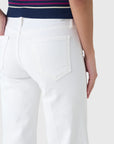 A person is shown from the waist down wearing Mother’s The Roller Fray high-waisted wide leg white jeans, paired with a navy top with pink stripes. One hand rests by their side and the jeans’ back pocket is visible.