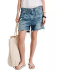 A person in a white shirt and Denimist's BROOKE SHORT distressed denim shorts, holding a beige tote bag. They wear white flip-flops and a brown wristband, highlighting the casual five-pocket style.